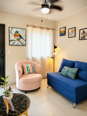 Ein Wohnzimmer mit Deckenventilator, einem blauen Sofa, einem rosa Sessel, einem schwarzen Couchtisch und einer Stehlampe.
