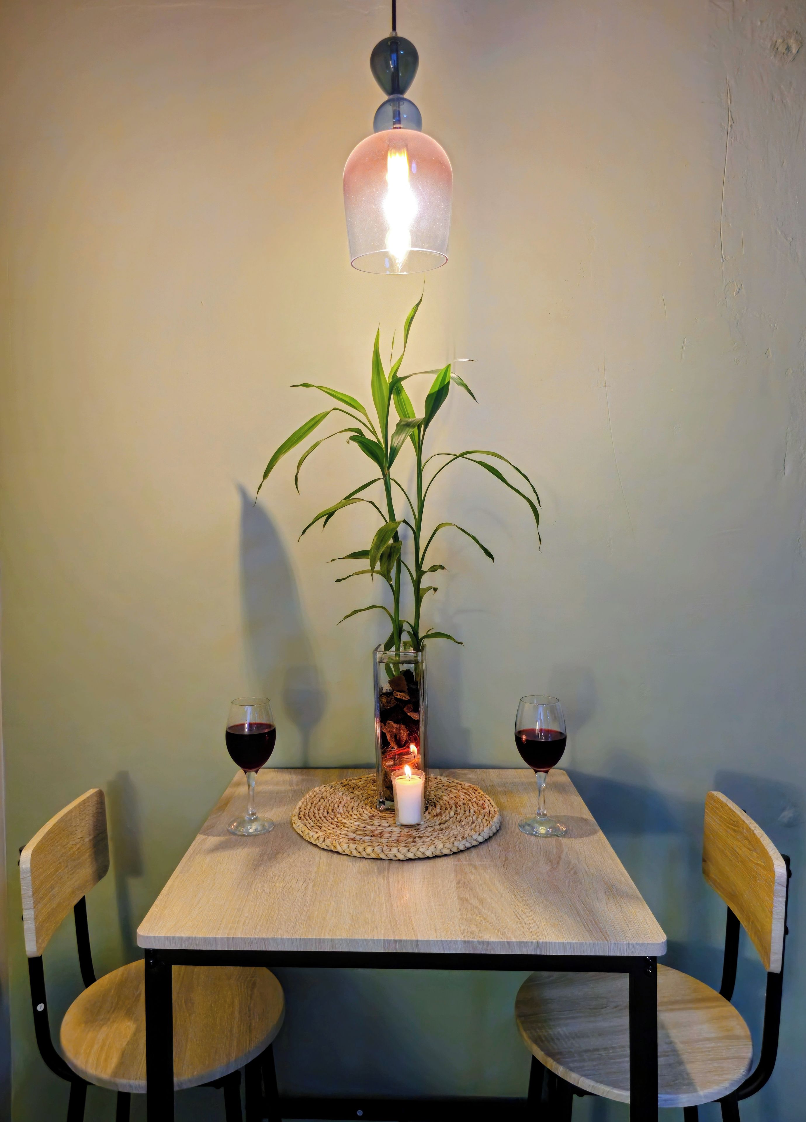 A dining table with two chairs, a candle, a glass vase with a plant, and two glasses of red wine.