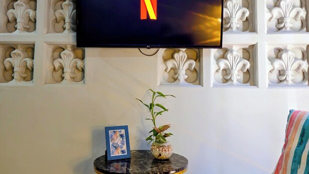 Una TV a schermo piatto montata su una parete decorata con motivi a giglio, un tavolino con una pianta e una cornice, e un cuscino a righe su un divano.