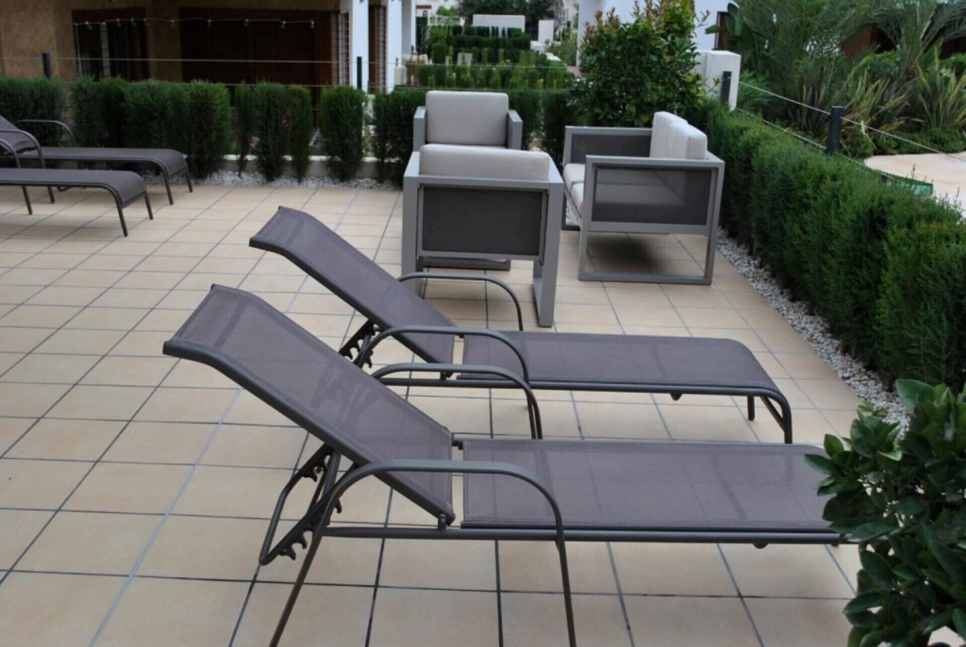 Outdoor lounge chairs and tables arranged on a tiled patio with a view of a building and greenery.