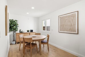 A dining area with a wooden table and chairs, a potted plant, a kitchen area with cabinets, and a framed artwork on the wall.