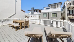 En tagterrasse med et træbord og loungesenge, et hvidt plankeværk og tilstødende bygninger.