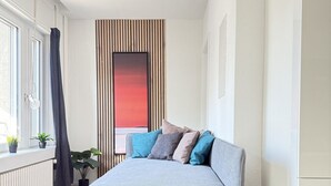 A modern bedroom with a grey sofa bed, a large window with curtains, a radiator, and a potted plant. - Stylish studio apartment near the Olma trade fair (St. Gallen)