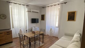Een woonkamer met een bank, een eettafel met stoelen, een aan de muur bevestigde televisie en een airconditioning.