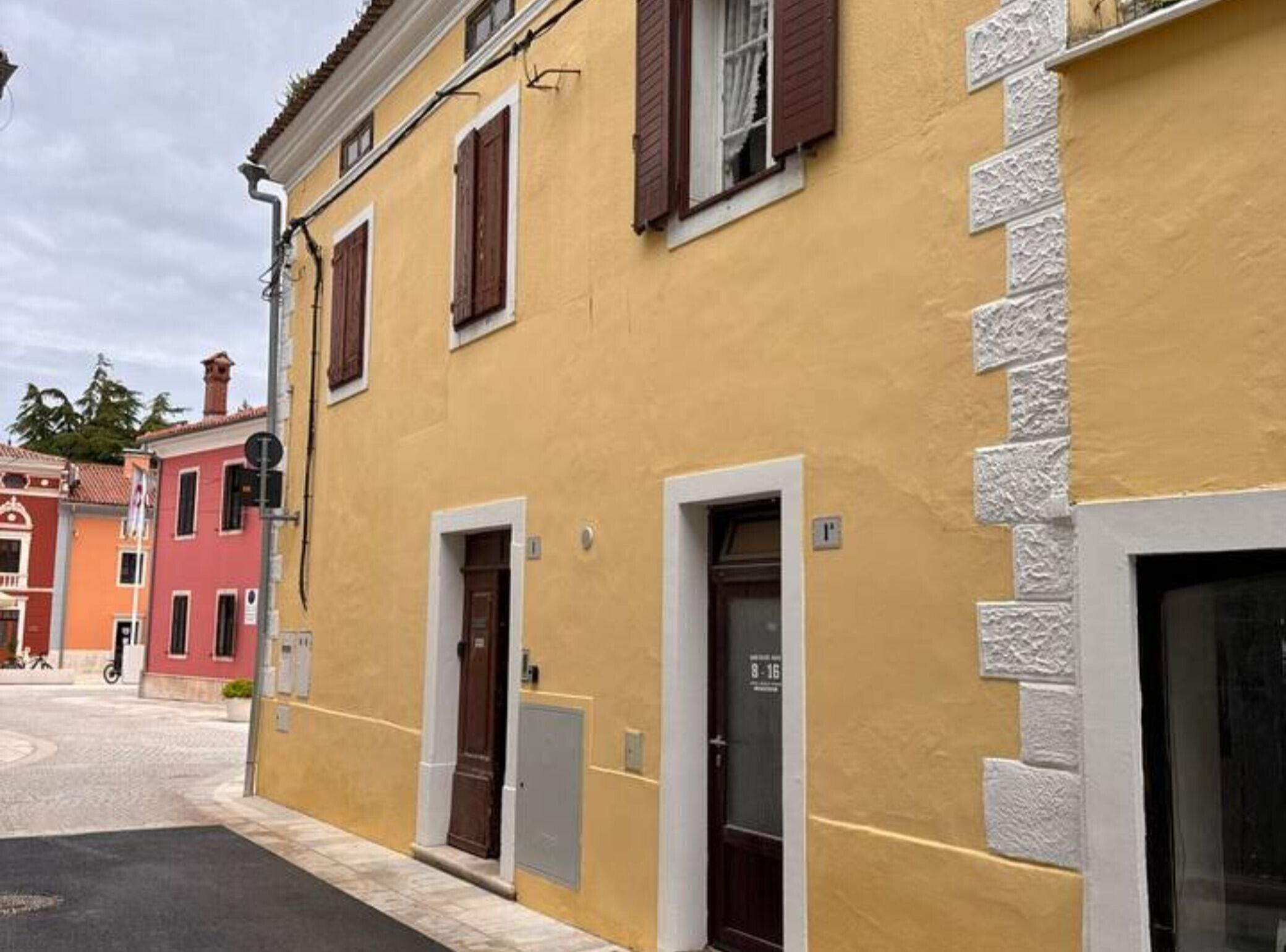 Un bâtiment jaune avec deux portes, une fenêtre avec des volets et une petite enseigne au-dessus de la porte.