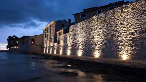 Een stenen muur met daarlangs een gebouw, beide verlicht door kunstlicht.