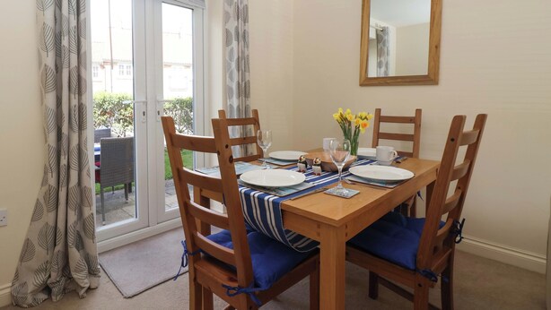 A dining area with a table set for four, wooden chairs with blue cushions, a vase with yellow flowers, and a mirror on the wall. - Sea Glass Cottage (Filey)