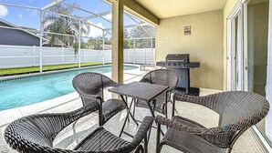 Um alpendre coberto com móveis de vime, churrasqueira e vista para uma piscina.