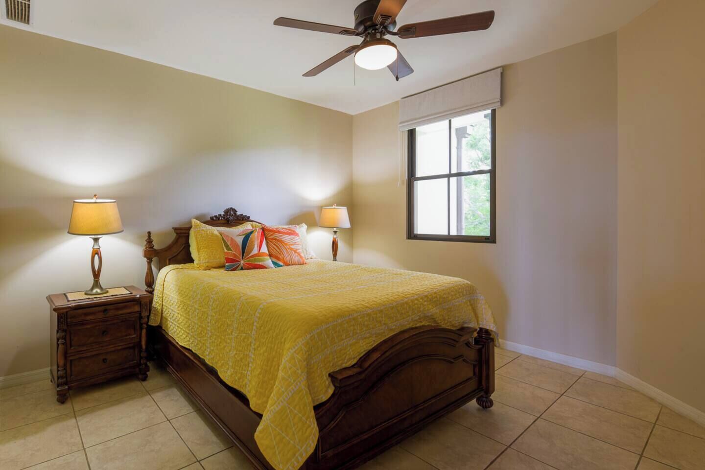 A bedroom with a bed, two bedside tables with lamps, a ceiling fan, a window, and a view of greenery outside.