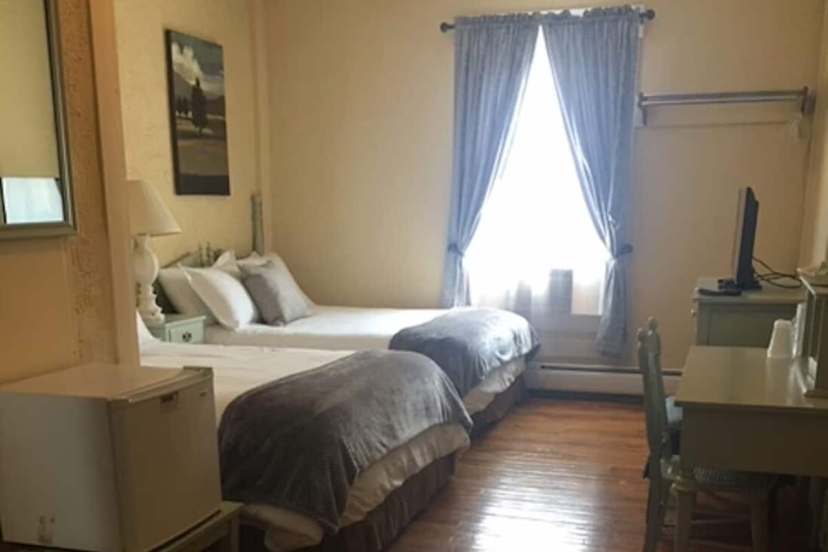 A hotel room with two beds, a desk, a chair, a small table, a lamp, a picture on the wall, and a window with curtains.