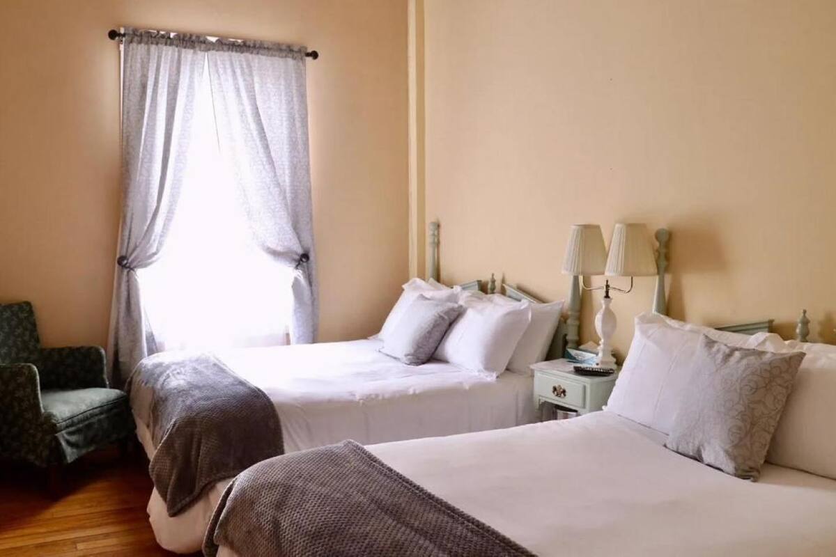 A hotel room with two beds, a chair, a nightstand with lamps, and a window with curtains.