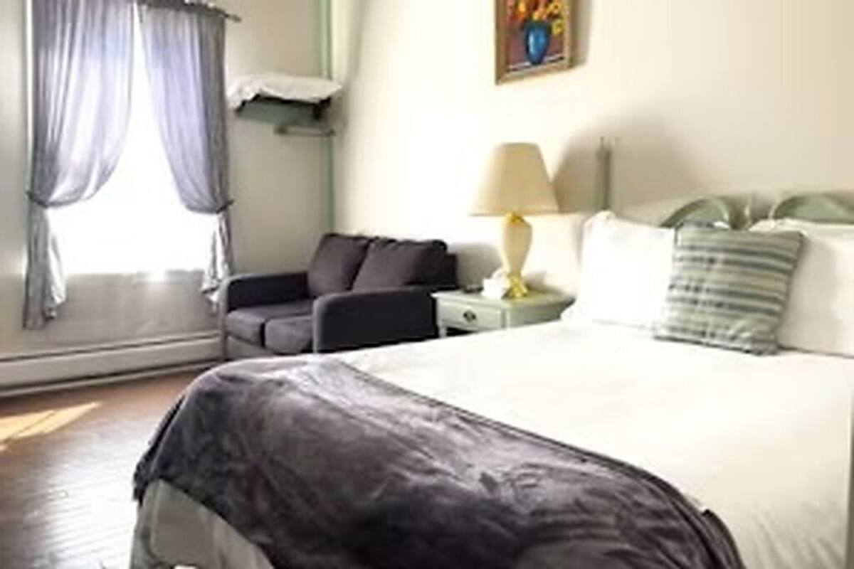 A hotel room with a bed, a nightstand, a lamp, a window with curtains, and a painting on the wall.