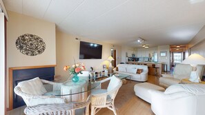 A living room with a glass coffee table, wicker chairs, a sofa, a TV, and a kitchen in the background. - Waters Edge - A 3 bedroom, 2 bathroom, condo located right on the beach! (Brigantine)