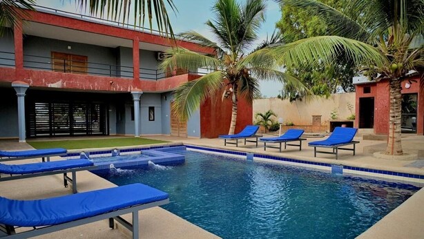 A swimming pool with blue lounge chairs, palm trees, and a building with a red and grey facade. - la villa du parc (Niaga)