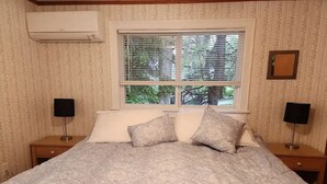 A bedroom with a bed, two bedside tables, a window with blinds, an air conditioner, and a wall-mounted TV. - Charming 2-bedroom house in peaceful Ogunquit with AC and screened porch. (Ogunquit)