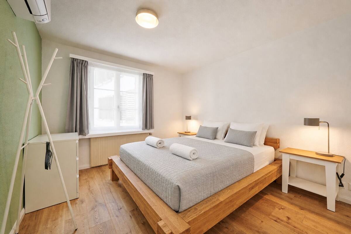 A modern bedroom with a wooden bed, bedside tables, a nightstand, a window with curtains, and a radiator.