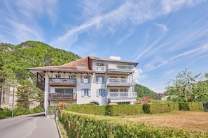 Un immeuble à plusieurs étages doté de balcons, une route en contrebas et un paysage vallonné en arrière-plan.