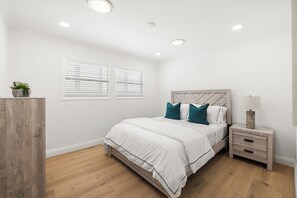 Ein Schlafzimmer mit einem Bett, Nachttischen, einer Lampe, einer Topfpflanze und einem Fenster mit Jalousien.
