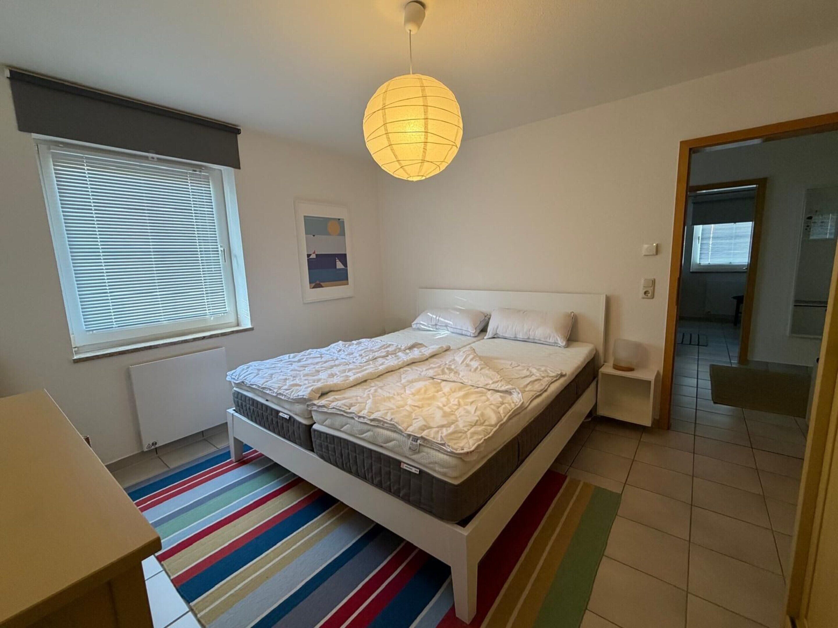 Une chambre à coucher comprenant un lit, une table de chevet, une fenêtre avec des stores, un tapis rayé aux couleurs vives et un luminaire suspendu.