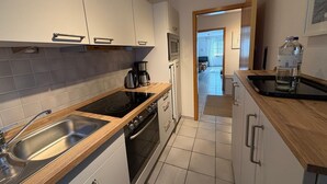Une cuisine moderne équipée d’une cuisinière, d’un évier et d’une machine à café. On aperçoit l’espace de vie à travers une porte ouverte.