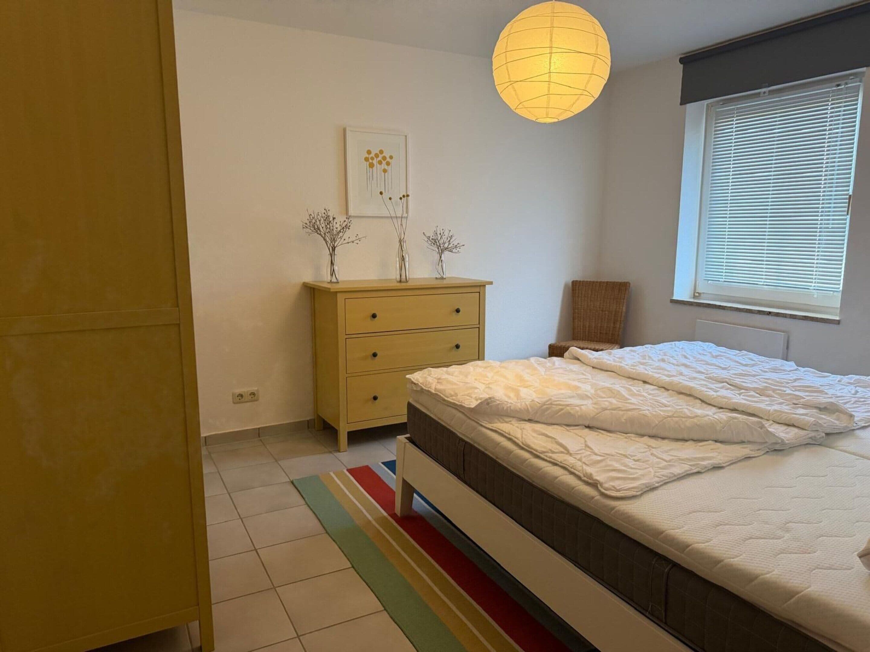 Une chambre à coucher comprenant un lit, une commode, une fenêtre avec des stores et un luminaire suspendu.