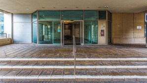 Entrada de un edificio con puertas de cristal, baranda metálica y suelo de baldosas.
