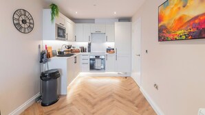 Una cocina moderna con armarios blancos, un microondas y una estufa. Hay un cubo de basura y un reloj en la pared.