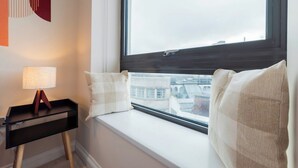 Una habitación con un asiento junto a la ventana, una lámpara y una obra de arte en la pared.