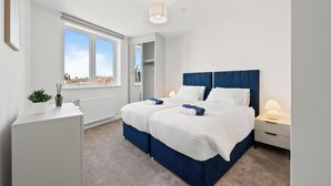 A bedroom with a large bed, a desk, a chair, a plant, and a window with a view. - Thamesview | Heathrow | 2 Bed (Staines-upon-Thames)