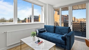 A modern living room with a blue sofa, a white coffee table, and a view of the city through large windows. - Thamesview | Heathrow | 2 Bed (Staines-upon-Thames)