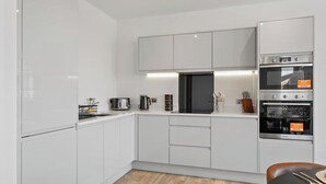 A modern kitchen with white cabinets, a black refrigerator, a double oven, and a wooden floor. - Thamesview | Heathrow | 2 Bed (Staines-upon-Thames)