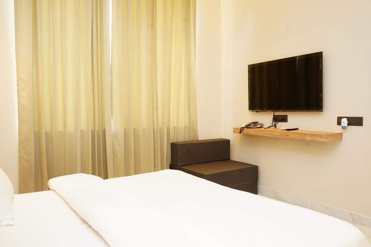 Quarto de hotel com cama, um pequeno espaço de estar, televisão montada e uma prateleira de madeira com um telefone.