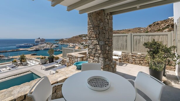 Eine Terrasse mit einer Steinsäule, weißen Stühlen, einem runden Tisch, einem Pool und Blick auf das Meer und den Hafen.