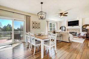 A spacious living room with a wooden floor, a dining table with white chairs, a sofa, a fireplace, a TV, and a ceiling fan.