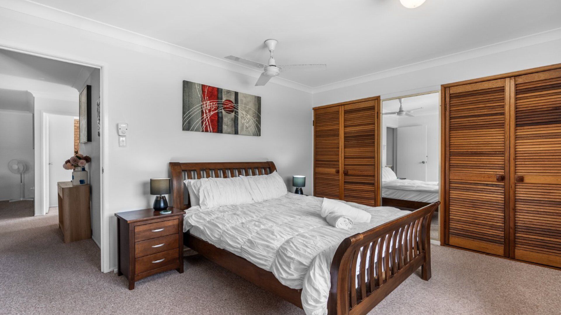 A bedroom with a wooden bed, bedside tables, a ceiling fan, a large mirror, and a wardrobe.