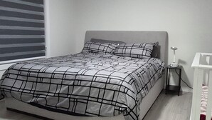 A bedroom with a bed, a nightstand, a crib, and a window with blinds. - Vibrant Homestead (Edmonton)