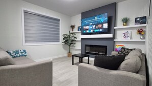 A living room with two sofas, a fireplace, a TV mounted above it, and a window with blinds. - Vibrant Homestead (Edmonton)