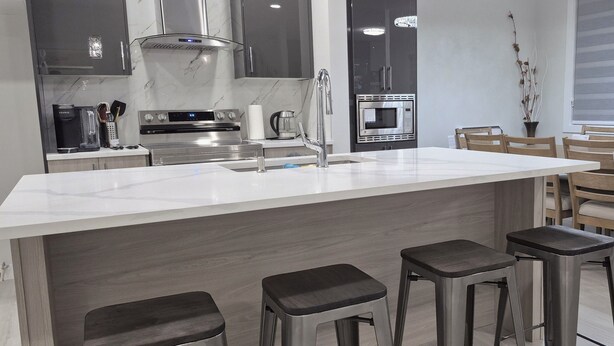 A modern kitchen with a central island, bar stools, and a dining area with wooden chairs. - Vibrant Homestead (Edmonton)