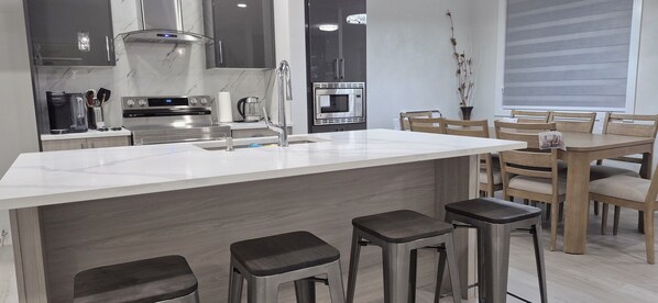 A modern kitchen with a central island, bar stools, and a dining area with wooden chairs. - Vibrant Homestead (Edmonton)