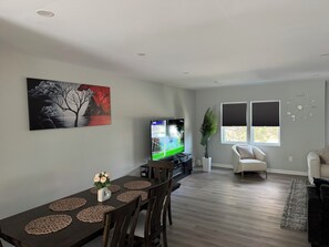 Ett modernt vardagsrum med en platt-TV, ett matbord med bordsunderlägg, en tavla, en krukväxt och en soffa.