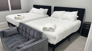 A hotel room with two beds, a grey upholstered chair, and a small bedside table. - Spacious 5-bedroom house in perfect Bowie with cool AC and gym (Bowie)