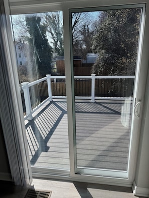 Vista a través de una puerta corrediza de vidrio a una terraza de madera con baranda blanca, que da a un patio trasero con árboles y una valla de madera.