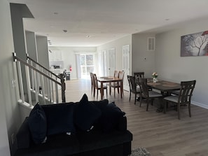 Una sala de estar moderna con un sofá oscuro, una zona de comedor con sillas de madera y una mesa, y una escalera que conduce a un piso superior.
