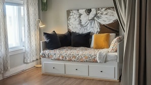 Une pièce avec un lit, une fenêtre avec des rideaux, un luminaire au plafond et une plante fixée au mur.