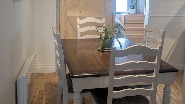 Un coin repas avec une table en bois et des chaises blanches, un luminaire au plafond, et une vue sur une pièce avec une fenêtre et des rideaux.