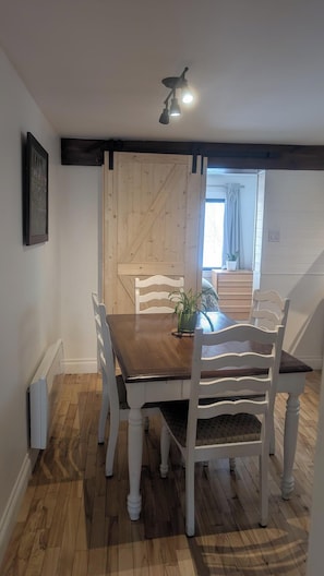 Un coin repas avec une table en bois et des chaises blanches, un luminaire au plafond, et une vue sur une pièce avec une fenêtre et des rideaux.