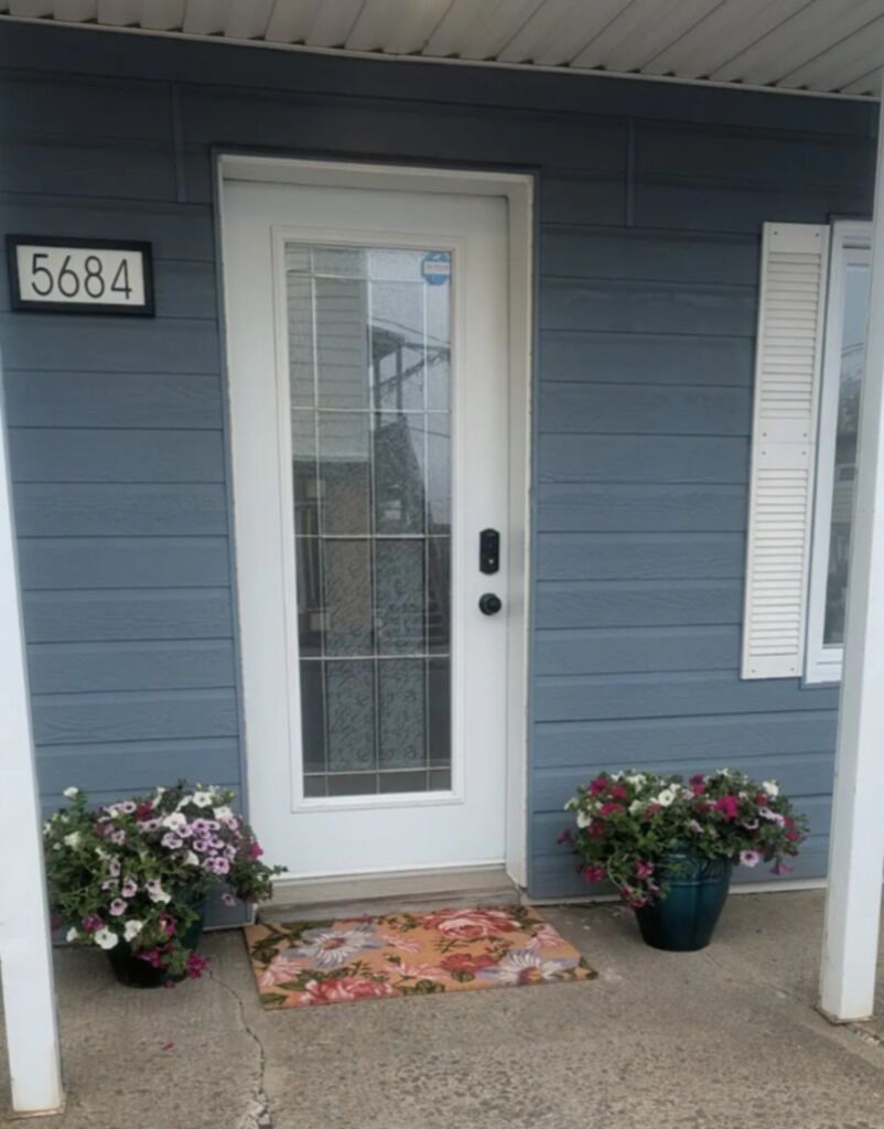 A front door with a number '5684' above it, two potted plants, and a floral mat on the concrete porch.