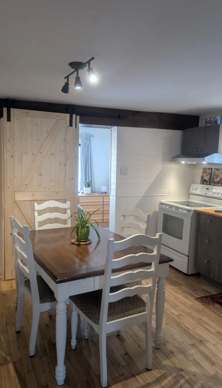 A dining area with a wooden table and white chairs, a kitchen with a stove and cabinets, and a wooden door.