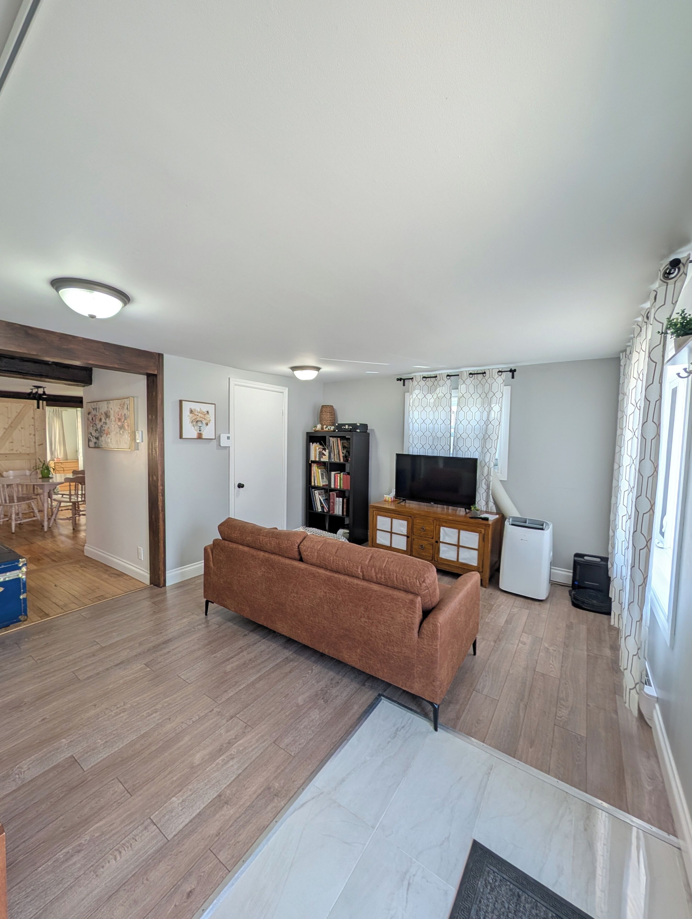 Una sala de estar con un sofá marrón, televisión, estantería y comedor con muebles de madera.