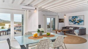 A modern living room with a glass dining table, a grey sofa, and a painting on the wall. - Kalafatis Stay With Private Pool (Mykonos)